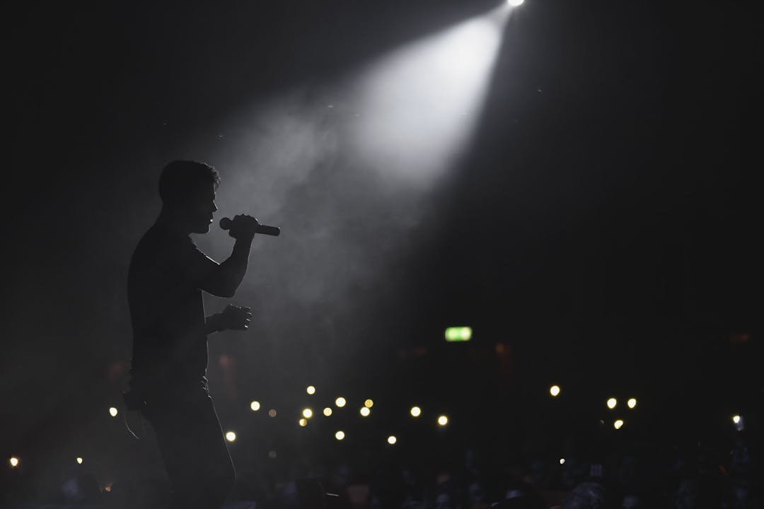Silhueta de cantor com microfone sob spotlight em palco, plateia ao fundo com luzes