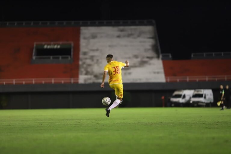 Jogador de futebol com camisa amarela em movimento de chute durante partida noturna em estadio