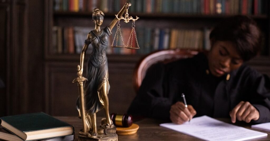 Estátua da Justiça com balança em primeiro plano e juíza assinando documento ao fundo
