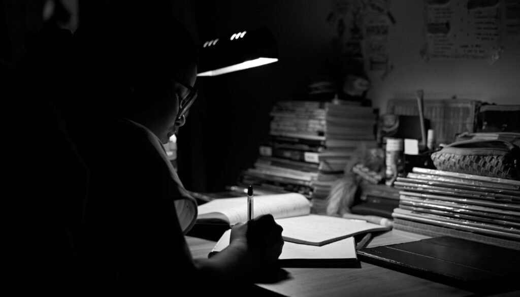 Estudante escreve em caderno sob a luz de uma luminária, cercado por pilhas de livros