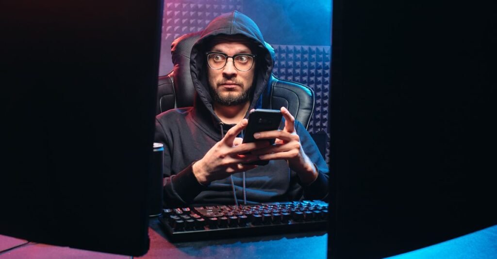 Homem de capuz usa smartphone em ambiente escuro diante de computador