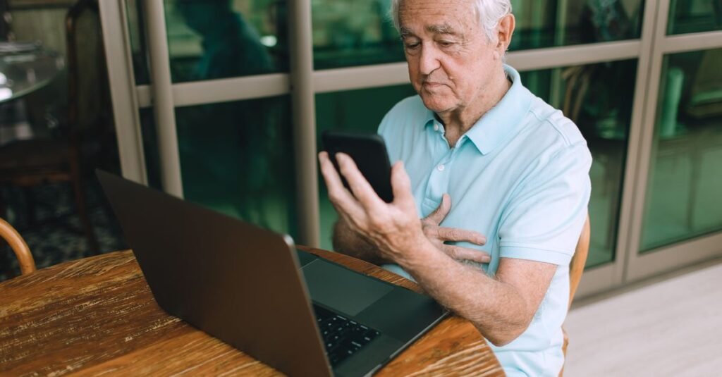 Homem idoso usando smartphone diante de um notebook sobre mesa de madeira