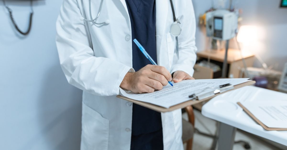 Médico de jaleco branco escreve em prancheta em consultório