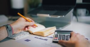 Mãos usando calculadora e anotando em caderno sobre mesa com documentos financeiros