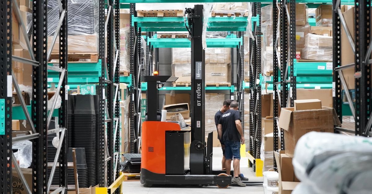 Trabalhadores e empilhadeira em corredor de galpão logístico com prateleiras