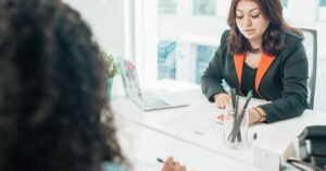 Duas pessoas em mesa de escritório durante entrevista de emprego, com currículo sobre a mesa