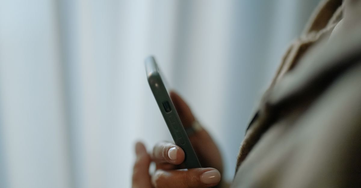 Close de uma mao segurando um telefone celular, em ambiente interno com fundo claro e desfocado
