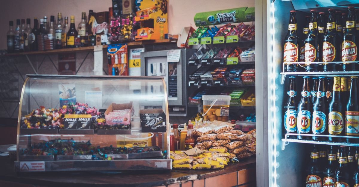 Balcão de loja de conveniência com doces, salgados e geladeira de bebidas ao fundo