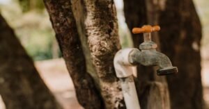 Torneira externa fechada, com registro de metal e cano branco, em área arborizada
