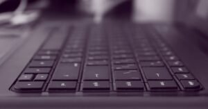 Teclado de computador preto em primeiro plano, com o restante da mesa desfocado