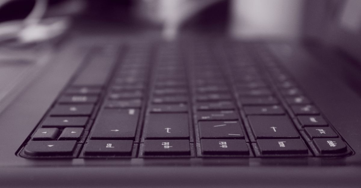 Teclado de computador preto em primeiro plano, com o restante da mesa desfocado