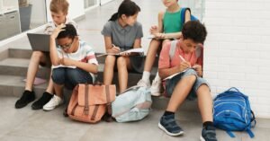 Grupo de adolescentes sentado na escada da escola escrevendo em cadernos, com mochilas ao lado
