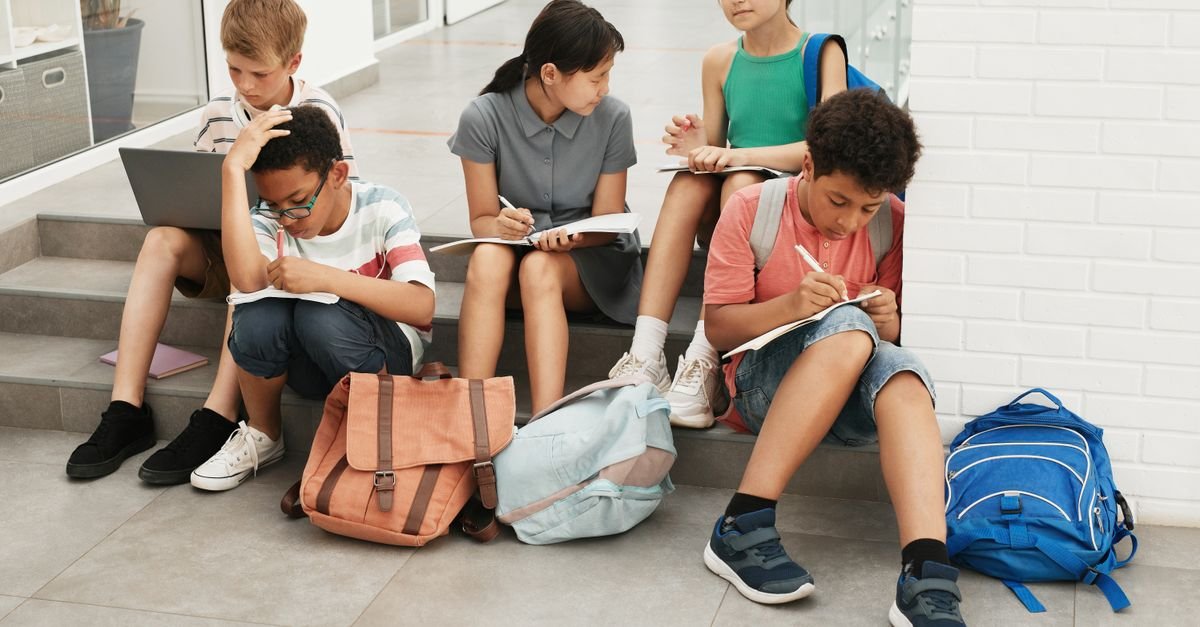 Grupo de adolescentes sentado na escada da escola escrevendo em cadernos, com mochilas ao lado