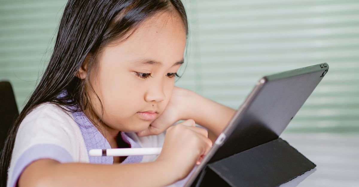 Criança concentrada usando caneta digital em um tablet apoiado sobre a mesa