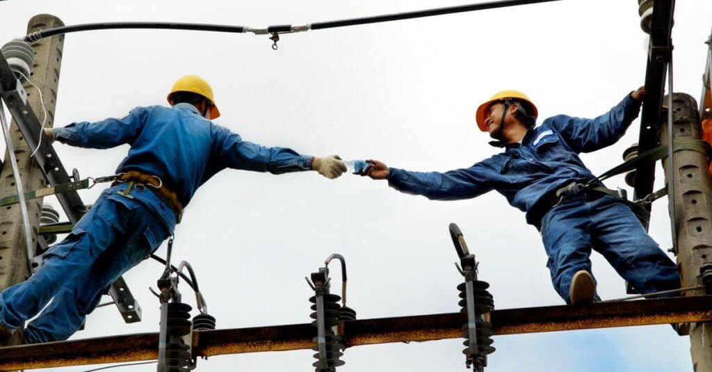 Dois eletricistas com capacete trabalham em rede de energia elétrica no alto de um poste
