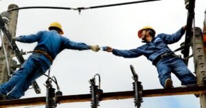 Dois eletricistas com capacete trabalham em rede de energia elétrica no alto de um poste