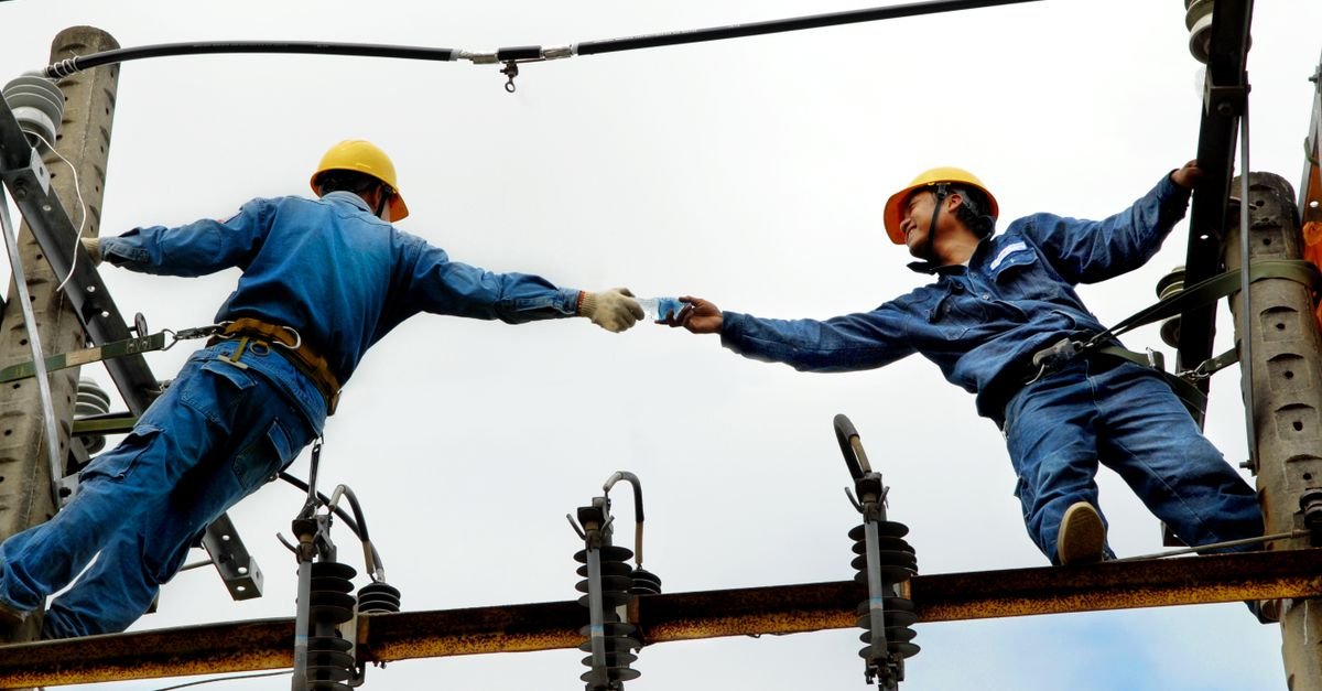 Dois eletricistas com capacete trabalham em rede de energia elétrica no alto de um poste
