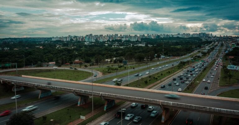 Carros em rodovia de pista dupla com viaduto e prédios ao fundo