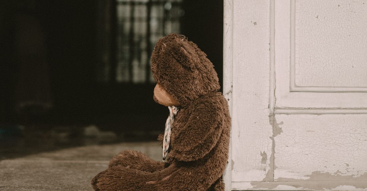 Urso de pelúcia sentado sozinho ao lado de uma porta, em imagem ilustrativa sobre proteção à infância