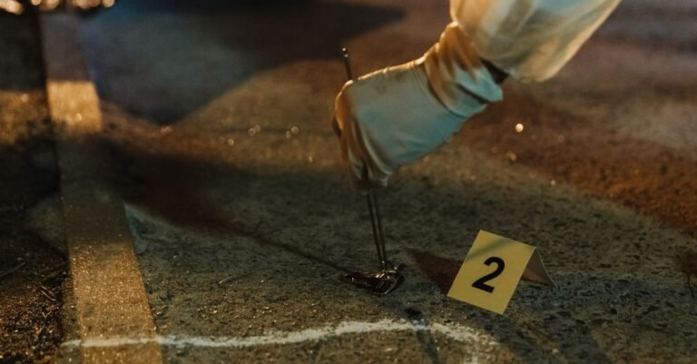 Perito com luvas recolhe uma cápsula deflagrada do asfalto ao lado de um marcador numérico amarelo em cena de crime à noite