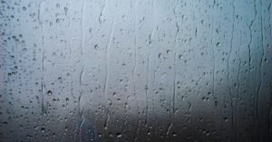 Vidro de janela coberto por gotas e escorridos de chuva, com fundo cinza desfocado