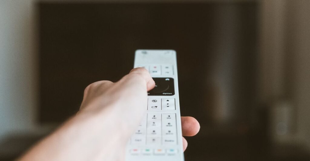 Mao aponta controle remoto branco para uma televisao desfocada ao fundo