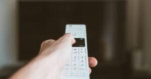 Mao aponta controle remoto branco para uma televisao desfocada ao fundo