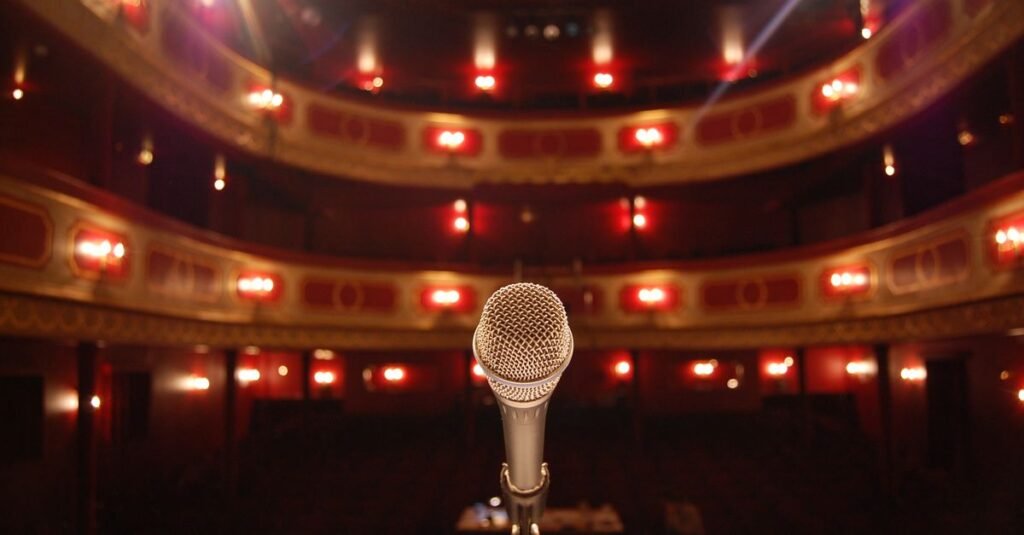 Microfone no centro de um palco de teatro com plateia e camarotes iluminados ao fundo
