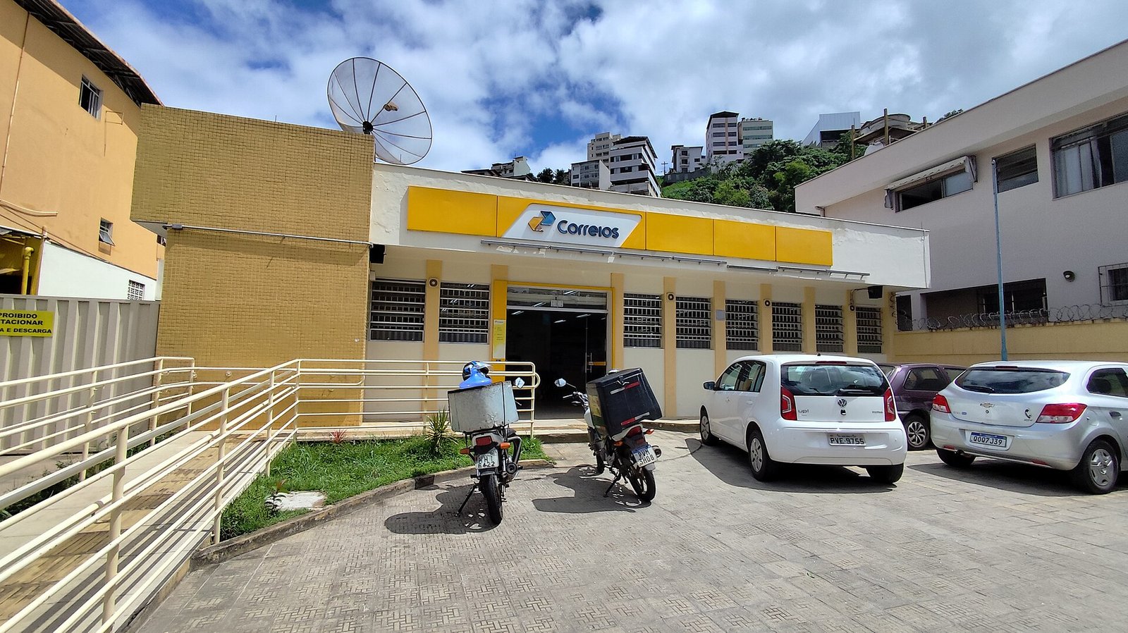 Fachada de agência dos Correios com letreiro amarelo e motos de entrega estacionadas