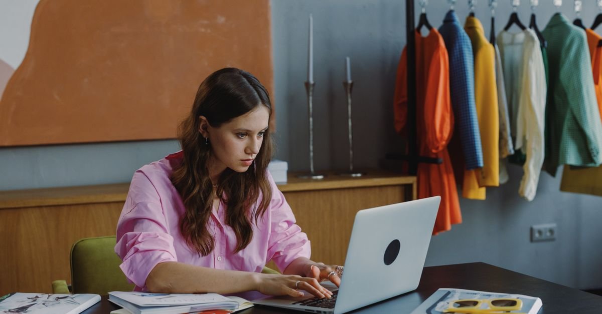 Empreendedora trabalha em notebook em ateliê com arara de roupas ao fundo