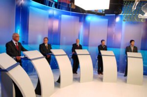 Candidatos posicionados em púlpitos durante debate em estúdio de televisão