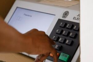 Mão de eleitor sobre o teclado numérico de uma urna eletrônica, com a tecla verde CONFIRMA à direita