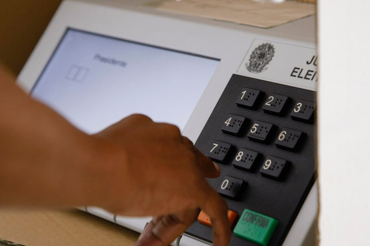 Mão de eleitor sobre o teclado numérico de uma urna eletrônica, com a tecla verde CONFIRMA à direita