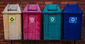 Quatro lixeiras de coleta seletiva identificadas como metal, plástico, vidro e papel, encostadas em muro de tijolos