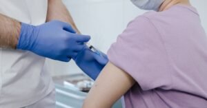 Profissional de saúde com luvas aplica dose de vacina no braço de um paciente em sala de atendimento
