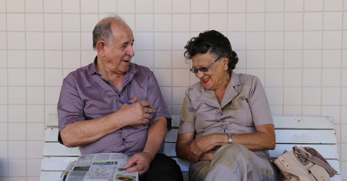 Casal de idosos conversa sentado em banco de praça com jornal
