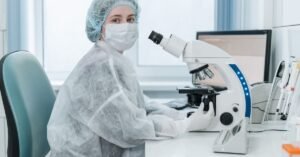 Técnica de laboratório com jaleco e touca observa amostras em microscópio