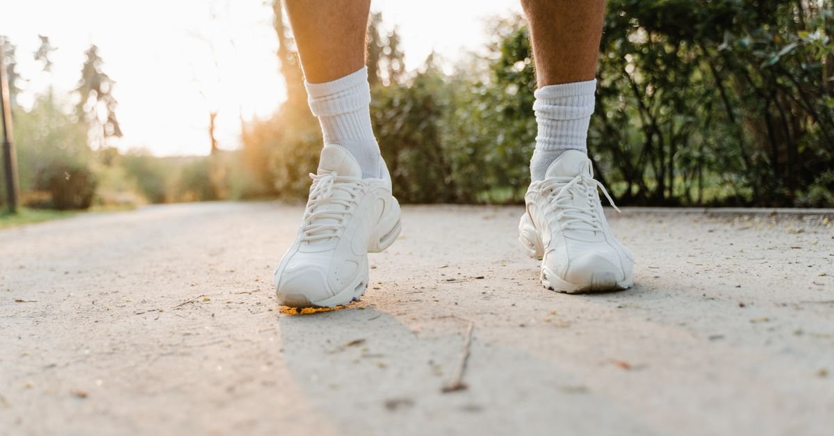 Close-up dos pes de uma pessoa caminhando de tenis branco por uma trilha de parque ao entardecer