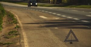 Triângulo de sinalização montado no acostamento de uma rodovia, com carro de faróis acesos ao fundo no fim de tarde