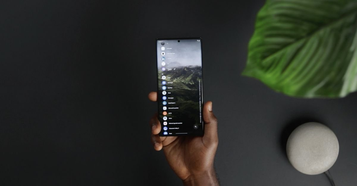 Mão segurando um smartphone com a lista de aplicativos na tela, sobre fundo escuro