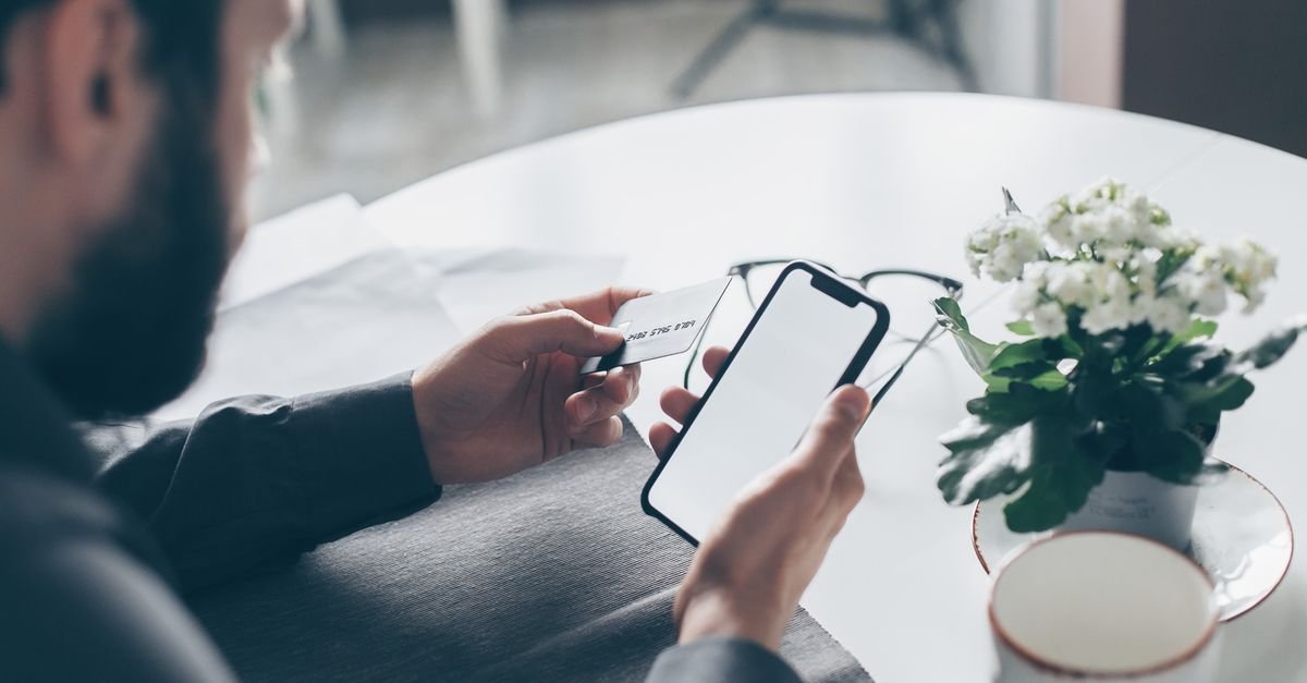Homem segura cartão bancário em uma das mãos e celular na outra durante operação financeira