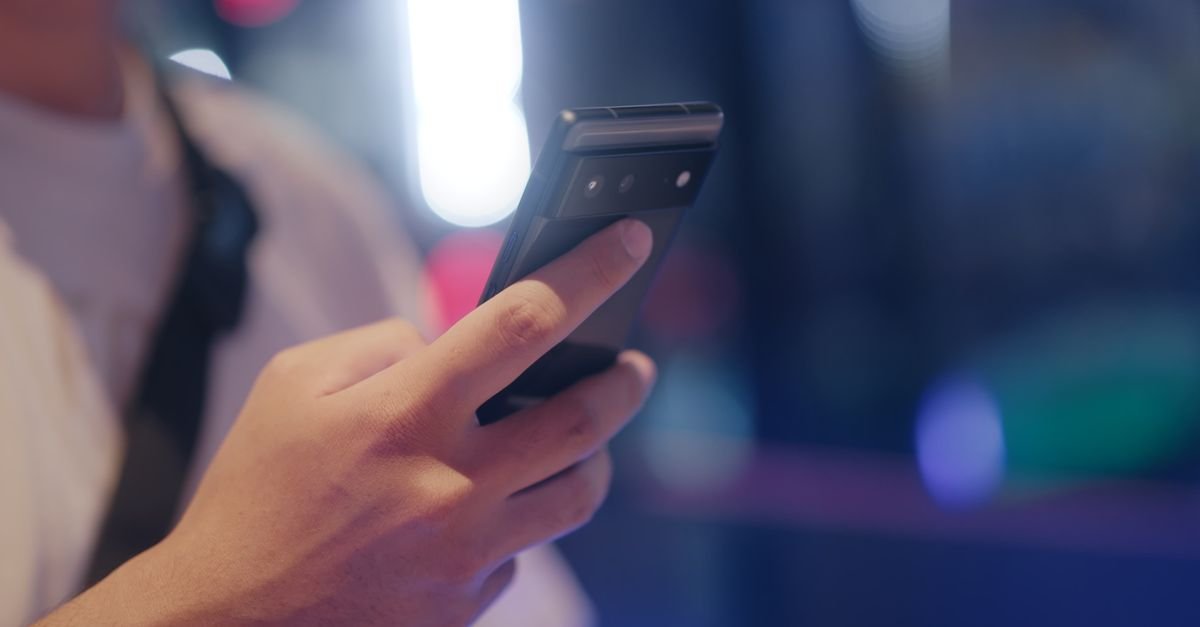 Mao de uma pessoa segurando um telefone celular em ambiente noturno com luzes desfocadas ao fundo