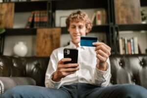 Homem sentado no sofá segura celular em uma das mãos e um cartão bancário na outra