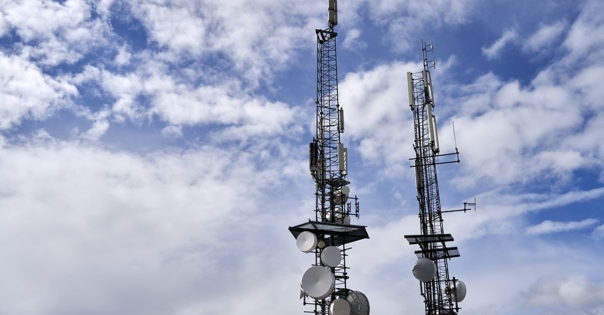 Duas torres de transmissão com antenas e parabólicas recortadas contra o céu azul com nuvens