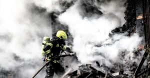Bombeiro combate incêndio com mangueira em meio à fumaça densa sobre escombros queimados