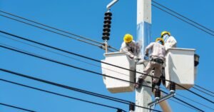 Dois eletricistas com capacete trabalham em cestas aéreas junto a um poste de concreto da rede elétrica