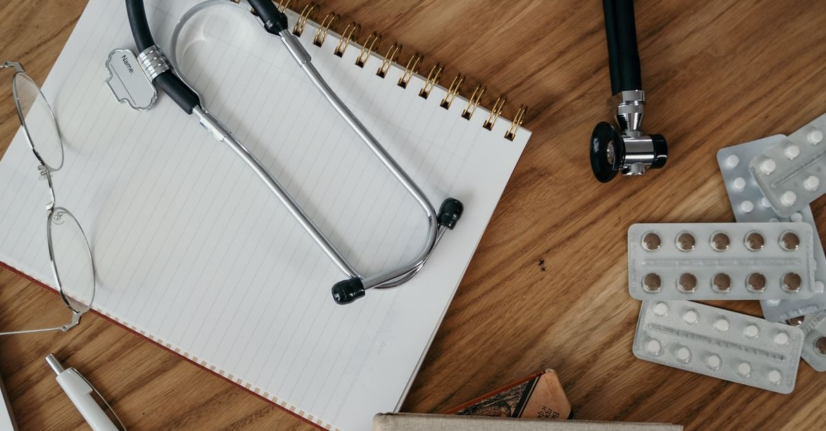 Estetoscópio sobre caderno, ao lado de óculos e cartelas de comprimidos