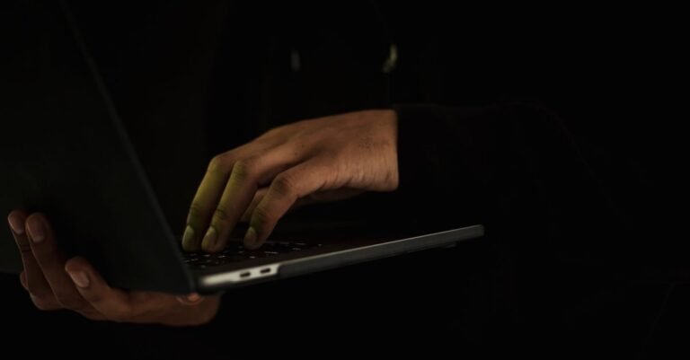 Mãos digitam em notebook em ambiente escuro, sem rosto visível
