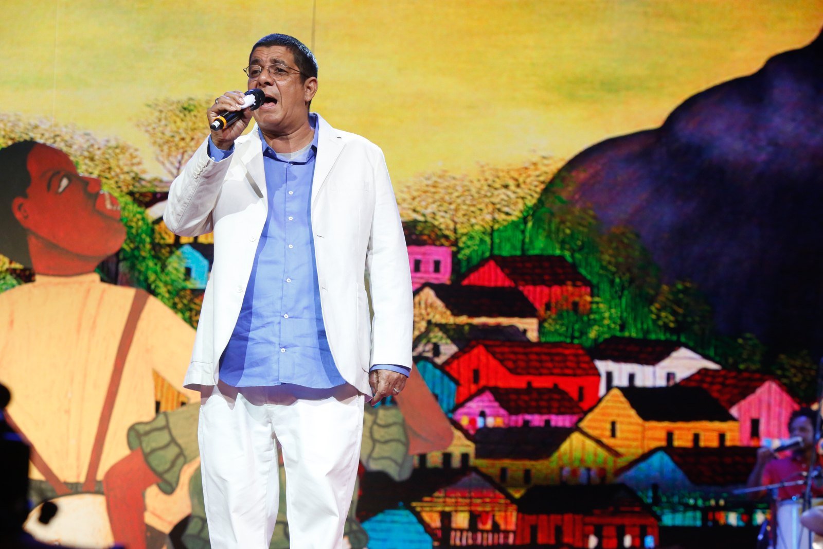 Zeca Pagodinho canta ao microfone em palco de premiação, de terno branco e camisa azul, diante de painel colorido