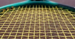 Close de raquete de tênis com encordoamento amarelo apoiada sobre quadra de saibro